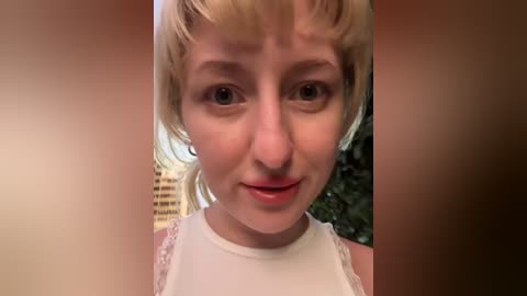 Media: Video of a young woman with short, blonde hair and fair skin, wearing a white lace-trimmed tank top, smiling with closed eyes. Background features blurred green foliage and a beige building.