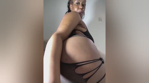 Video of a Black woman with medium skin tone wearing revealing black fishnet lingerie, posing seductively against a neutral wall, showing her ample buttocks and curves.