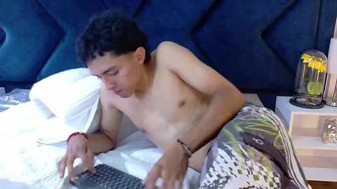 Video of a shirtless, slender Asian man with short black hair, lying on a bed, holding a phone, wearing gray shorts with a graphic design, in a modern bedroom with dark blue quilted headboard, white pillow, and nightstand with a lamp and books.