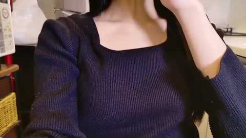 Video of a woman in a dark, textured sweater, seated indoors. She has long, dark hair, fair skin, and is partially obscured by her hand. The background features a kitchen with white cabinets and a red appliance.
