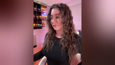 Video of a fair-skinned woman with long, curly brown hair wearing glasses and a black top, seated in a brightly lit room with shelves displaying colorful toys and a \"LEGO\" sign.