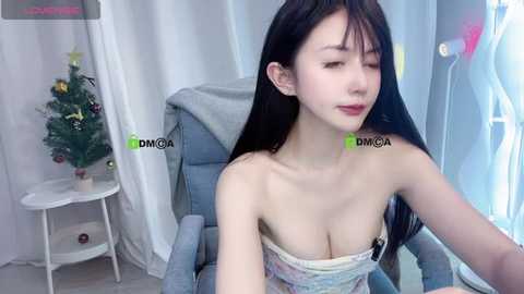 Video of an East Asian woman with long black hair, wearing a strapless floral dress, seated in a grey armchair. Background features a Christmas tree and white curtains.