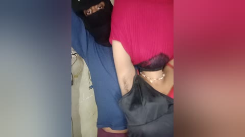 Video of a woman with fair skin, wearing a black mask, blue shirt, pink sweater, and black pants, holding a black bag, against a blurred background.