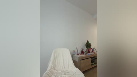 Video of a minimalist, white-walled room with a covered massage table in the foreground, and a wooden desk with red and green decor in the background.