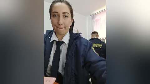 A video shows a young woman with light skin, dark hair pulled back, wearing a dark navy jacket over a white shirt and black tie. She holds a pink card. Background includes two uniformed security guards in dark jackets, one labeled \"BORDER GUARD.\