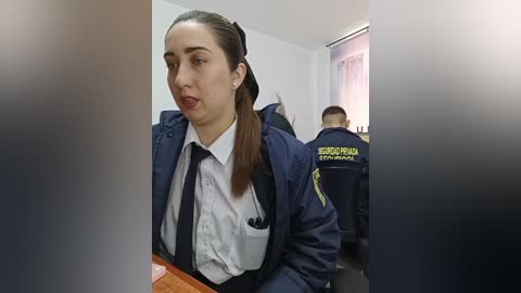 Video of a young woman with straight brown hair tied back, wearing a navy jacket, white shirt, and black tie, seated in a conference room with uniformed men in the background.