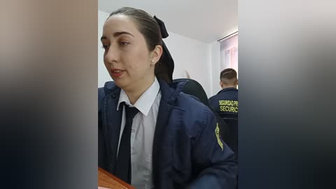 Video of a female police officer with straight, dark hair in a navy uniform, sitting in a dimly lit room with another officer in the background.