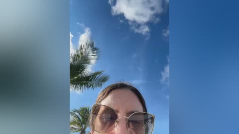 A video of a woman with light skin and brown hair, wearing large, gold-rimmed sunglasses, gazing upward at a bright blue sky with scattered clouds and a palm tree.