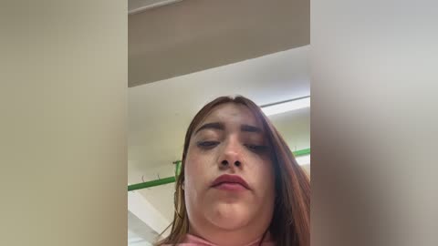 A video of a young Caucasian woman with fair skin and long brown hair, wearing a pink top, standing in a dimly lit room with beige walls and a green shelf.