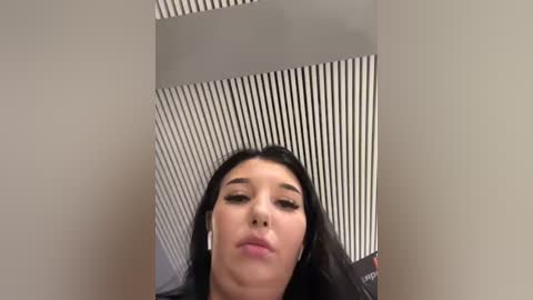 Video of a young woman with long dark hair, wearing black clothing, standing in front of a modern, white and black striped ceiling. She has a neutral expression, and the setting appears to be indoors.