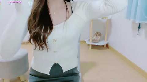 Video of a woman with long brown hair, wearing a white cardigan and gray skirt, adjusting her hair in a brightly lit, white-walled room with a small wooden table and chair.