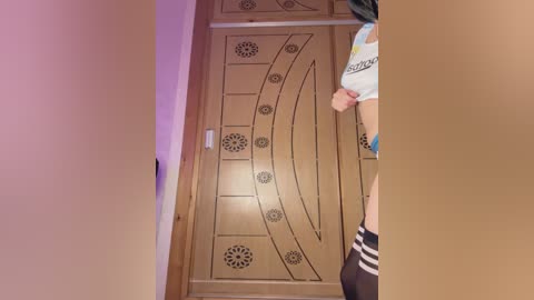 Video of a person, possibly male, standing in a doorway, wearing a white t-shirt and black shorts, with a wooden door featuring intricate circular patterns in the background.