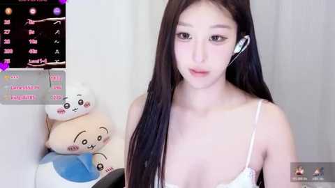 Video of a young East Asian woman with long black hair, wearing a white spaghetti-strap top, and a white earpiece, in a room with plush toys and a virtual reality headset.