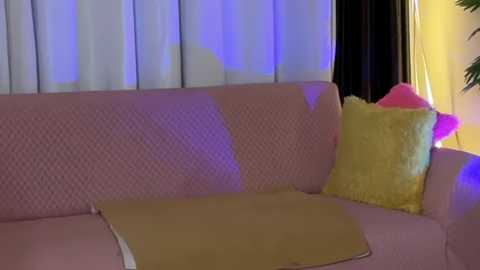A video shows a pink, patterned couch with a yellow throw and a bright yellow pillow, illuminated by purple lighting, in a cozy living room with white curtains.