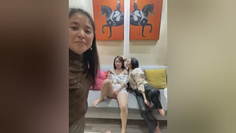 Video of two women with medium skin tones, one Asian, one Caucasian, wearing casual clothes, sitting on a couch, in a modern living room with orange horse paintings on the wall.