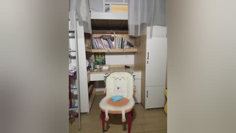 Video of a small, cluttered room with a white desk, a white chair with red legs, and a white bed with gray curtains. Shelves hold books and clothes. A metal ladder leans against the wall.