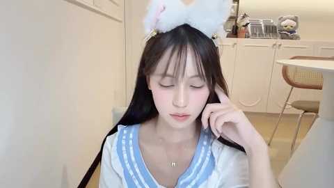 Video of an Asian woman with long black hair, wearing a white sailor dress with blue accents, and a fluffy white cat headband. She's sitting indoors, with white cabinets and a table in the background.