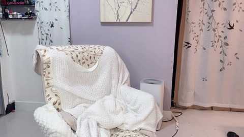 Video of a cozy, pastel-colored room with a white armchair draped in a knitted blanket, framed botanical artwork, light lavender walls, and beige curtains with leaf patterns.