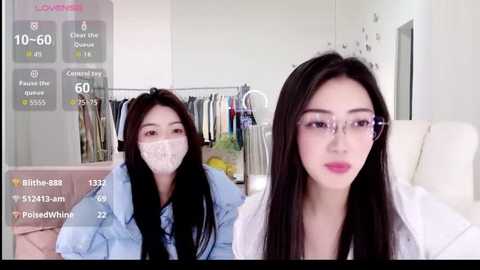Video of a young Asian woman with long black hair, wearing glasses, a white mask, and a light blue denim jacket, sitting on a white sofa. A virtual overlay shows social media stats.
