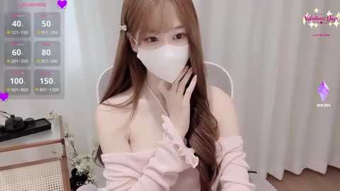 A video of an Asian woman with long brown hair, wearing a white face mask, pink off-shoulder top, and sitting at a desk in a room with white curtains.