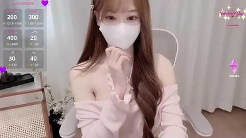 A video of an East Asian woman with long brown hair, wearing a white face mask, off-shoulder pastel pink blouse, and white headphones, seated indoors with a digital camera overlay.