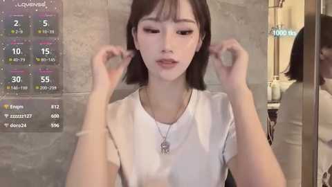 Video of a young East Asian woman with short black hair, wearing a white top, adjusting her hair in a bathroom mirror. Background shows tiled walls and a digital clock displaying 10:00.