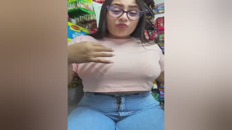 Video of a plus-sized woman with long dark hair, wearing glasses and a pink crop top, seated in a colorful candy store, making a pouty face.