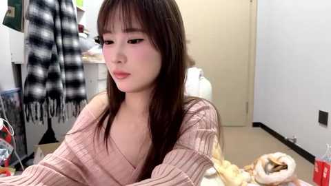 Video of a young Asian woman with straight brown hair, wearing a pink sweater, sitting in a cluttered room. A cat is on her lap.