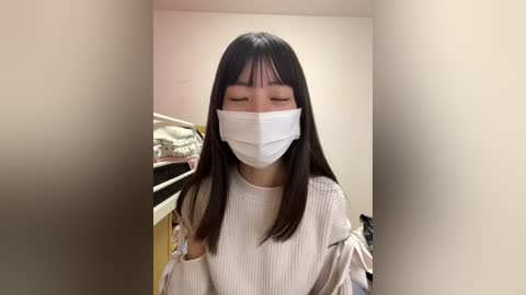 Media: Video of an East Asian woman with long black hair and fair skin, wearing a white surgical mask, gray sweater, and standing indoors in a dimly lit room with a bed and nightstand in the background.