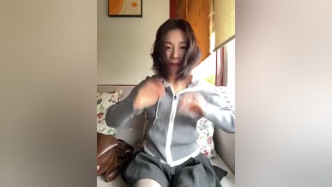 Video of a young Asian woman with shoulder-length brown hair, wearing a gray hoodie and black skirt, sitting on a floral-patterned couch in a sunlit room, adjusting her hoodie.