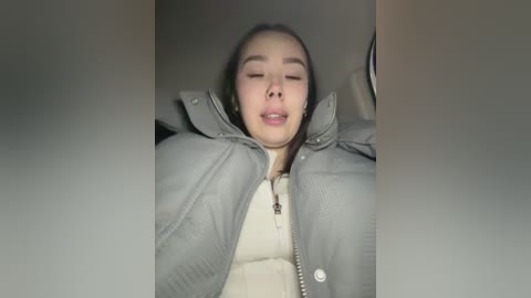 A video of a young Asian woman with closed eyes, resting her head back, wearing a gray puffer jacket and beige sweater, inside a car.