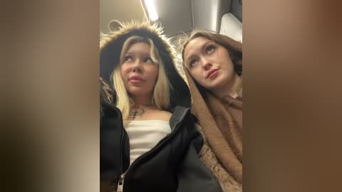 Media: Video of two young women in winter coats, one with blonde hair, one with brown hair, in an indoor setting, likely a bus or train.