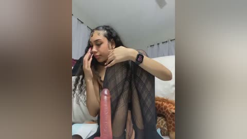 Video of a woman with long, wavy hair, wearing a white top and black sheer leggings, sitting on a bed, holding a large, erect dildo.