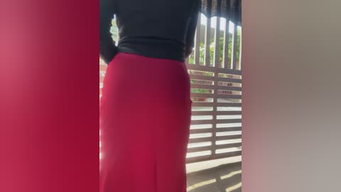 Video of a woman standing outdoors, wearing a black long-sleeve top and a vibrant red skirt, with a wooden fence and green foliage in the background.