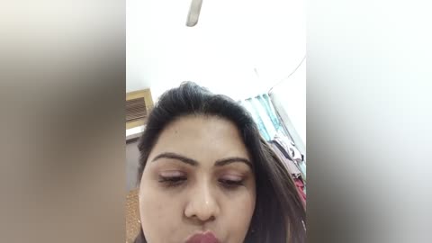 A video of a young woman with medium skin tone, dark hair, and light makeup, wearing a black top, captured from a low angle. Background features a white ceiling with a fan, a partially visible window, and a colorful blanket.