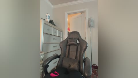 Video of a cluttered bedroom with a grey gaming chair, a silver dresser, a pink item on the chair, and a door open to a brightly lit room.