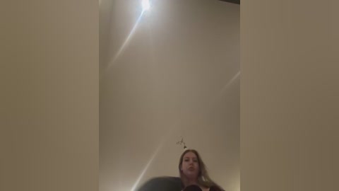 A video captures a woman with light skin and long, straight brown hair, partially visible from a low angle, standing in a dimly lit, beige-tiled hallway with a bright spotlight above her.