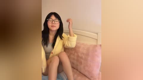 Video of a young Asian woman with long black hair, wearing glasses, a yellow hoodie, and grey top, sitting on a bed with a pink cushion and beige walls.