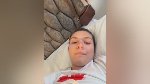 Media: Video of a young Caucasian boy with light skin and short brown hair, lying on a bed with a stone-textured headboard, wearing a gray shirt with a red graphic, and a brown leather bag.
