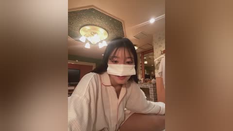 A video of an Asian woman with long black hair, wearing a white robe, surgical mask, and a bandage on her cheek, seated indoors with a chandelier, green walls, and a kitchen in the background.