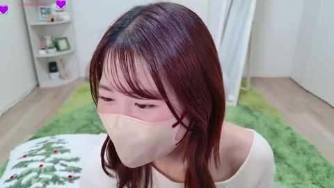 Video of an Asian woman with long, straight, dark brown hair, wearing a white mask, pink glasses, and a white top, standing in a brightly lit room with a green rug and white furniture.