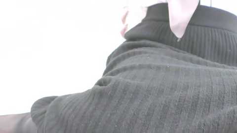 A close-up video of a person wearing a black, ribbed sweater with a white collar, lying on a bed. The background is plain white, emphasizing the texture of the sweater.