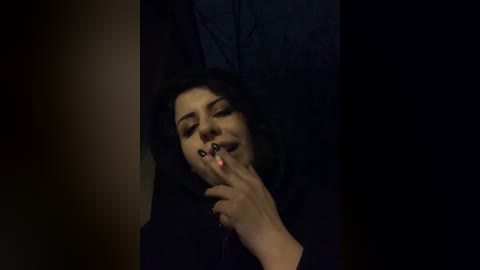 A video of a woman with dark hair, wearing a black dress, smoking a cigarette and blowing smoke rings, with a dim, dark background.