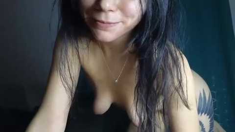 A video of a topless, light-skinned woman with long, dark hair and a tattoo on her right arm. She has a small, round breast and a thin necklace. The background is dark, possibly a dimly lit room.