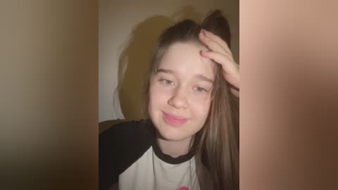 A video of a young Caucasian girl with light skin, brown hair in a high ponytail, and a smile. She wears a black and white t-shirt. Background is beige and slightly blurred.