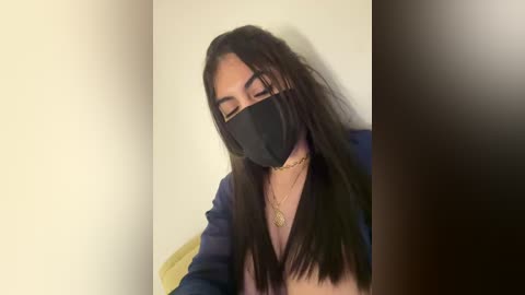 Media: Video of a young woman with long black hair, wearing a black face mask and a blue jacket, seated in a dimly lit room with a beige wall and a yellow object in the background.
