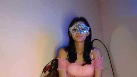 Video of a young woman with long black hair, wearing a pink off-shoulder top and a glittery masquerade mask. Background features a plain white wall and a black cable.
