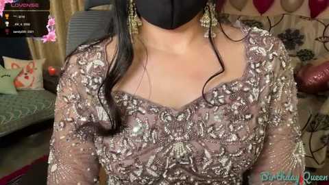 A video of a woman in a luxurious, ornate, brown gown adorned with intricate embroidery and sequins, wearing a black face mask, in a festive room with red balloons, cushions, and a bed.