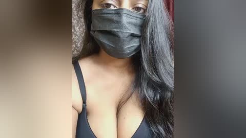 Video of a Black woman with medium skin tone, long dark hair, wearing a black face mask, revealing cleavage in a black bra. Background is blurred, possibly indoors.