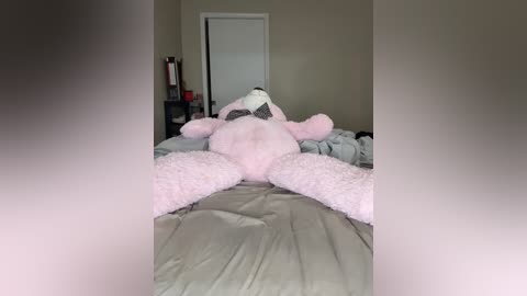 Video of a large, fluffy pink teddy bear with black bra, lying on a bed with grey sheets, in a dimly lit, beige-walled room.